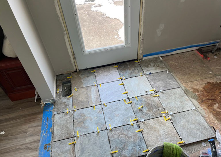 laid-slate-floor-repairs Newly laid slate tiles on a floor near a door, with spacers in place for grout. Adjacent area shows unfinished flooring and tools.
