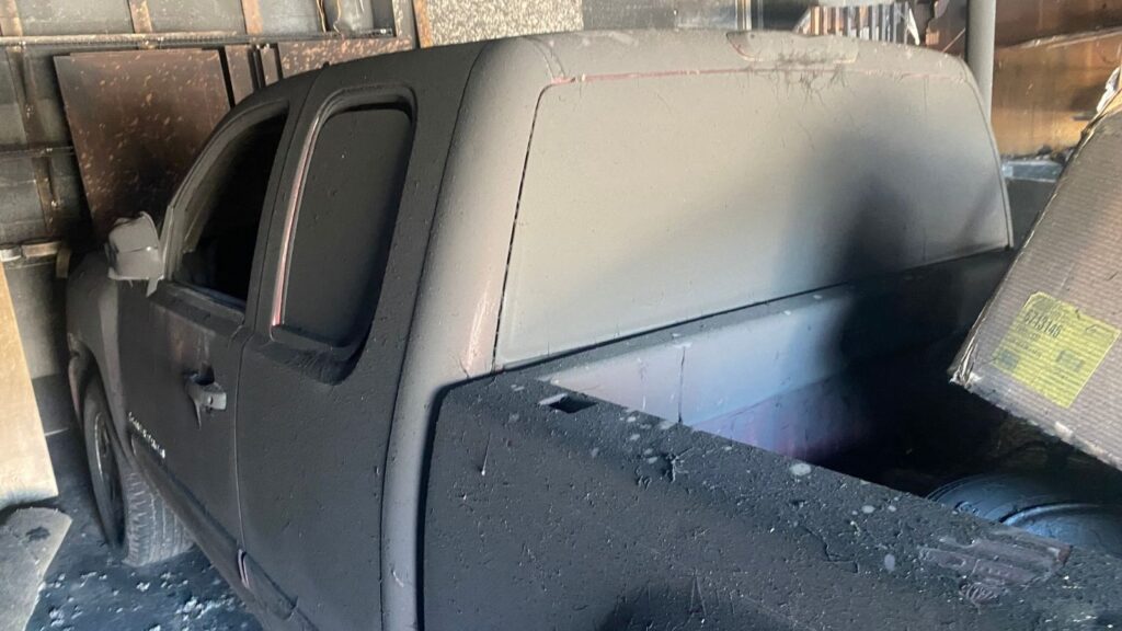A blackened pickup truck covered in soot and ash, parked in a garage with charred walls, indicating a fire incident.