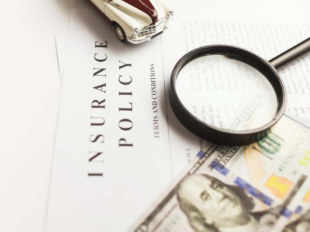 A vintage toy car rests on an insurance policy document, magnifying glass and cash bills scattered around, symbolizing financial review.
